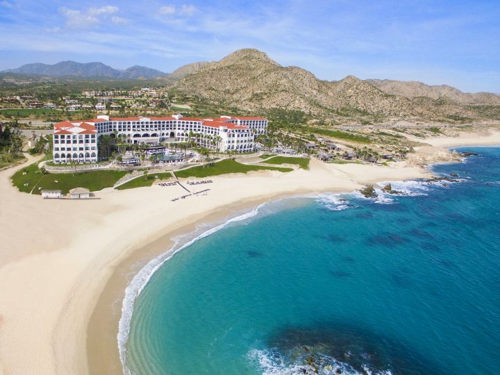 Aerial view of Hilton Los Cabos Beach & Golf Resort. Beach and coastline in the foreground. Hotel and mountains in the background.