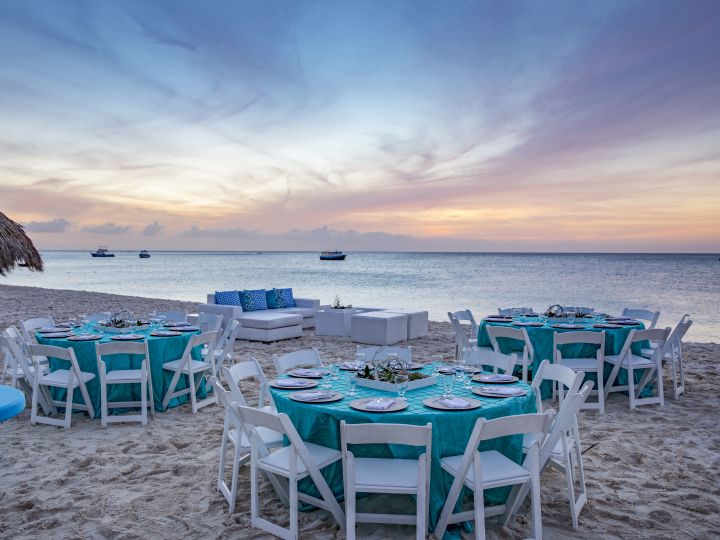 On-Site Beach Event at Sunset With Lounge Seating and Place Settings on Dining Tables With White Chairs