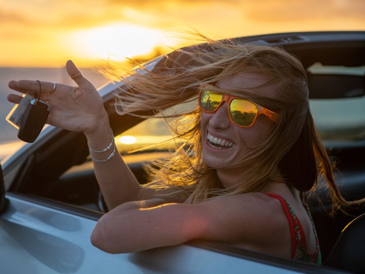 Woman in sunglasses holding car keys leaning out car with sunset in background