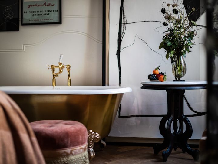 Bathroom with stand-alone gold bathtub, pink velvet chair, and side table holding a fruit plate.