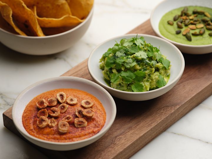 Puulauta, jossa on kulhoja punaista dipiä ja pähkinöitä, guacamolea ja vihreää salsaa siemenineen, kulhon vieressä