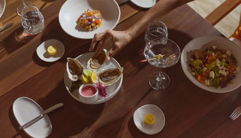 Woman's hand reaching for an oyster on a table set with a selection of fresh dishes and drinks
