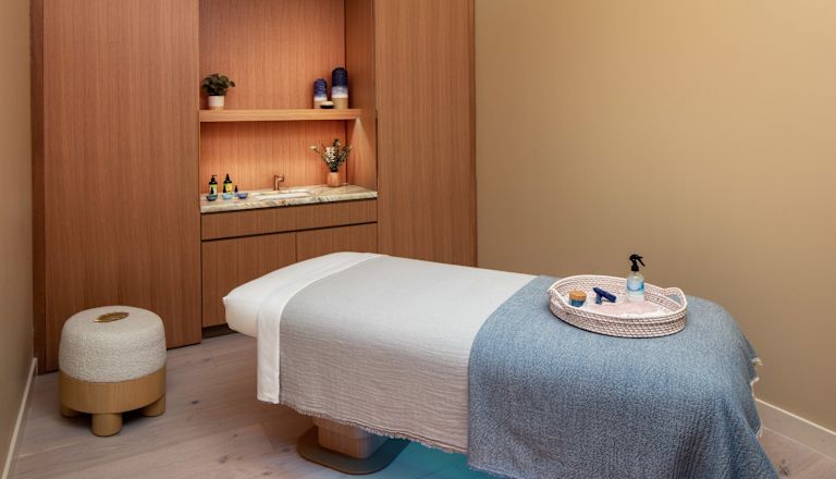 Camera de tratament spa cu masă de masaj, iluminare pentru atmosferă și produse într-o tavă împletită.
