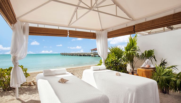 Two massage tables set up under a shelter with azure sea and a jetty in the background.