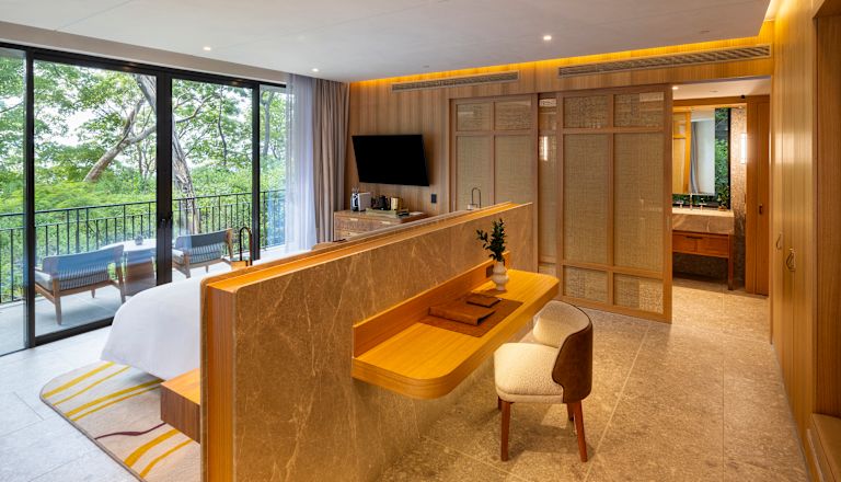 Dormitorio de la suite Pacifico, escritorio, cama, televisor, vista desde la ventana del balcón y los árboles