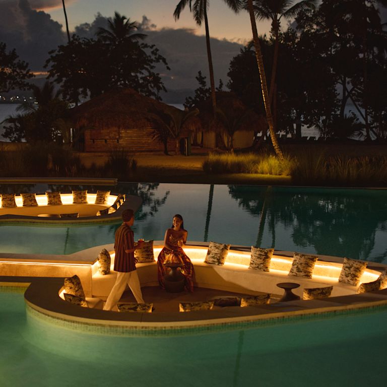 Two people in a softly lit sunken lounge surrounded by a pool at night, with warm accent lighting reflecting on the water and palm trees silhouetted against the dark sky.
