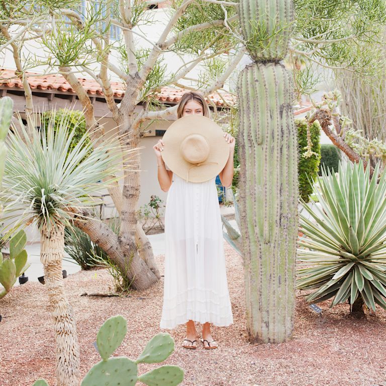 Woman holding hat in front of face, standing by cactus