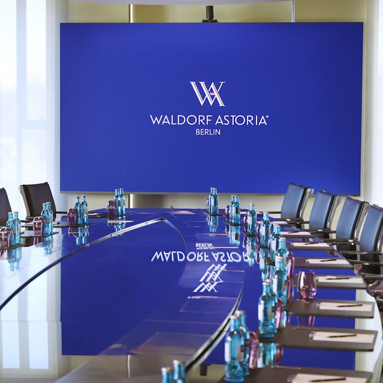 meeting room table with place settings, water bottles, blue screen with Waldorf Astoria Berlin text and logo