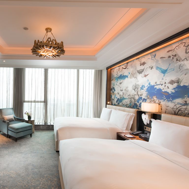 Deluxe room with two queen beds, soft chair, wardrobe, TV, work desk, and large art displayed above beds