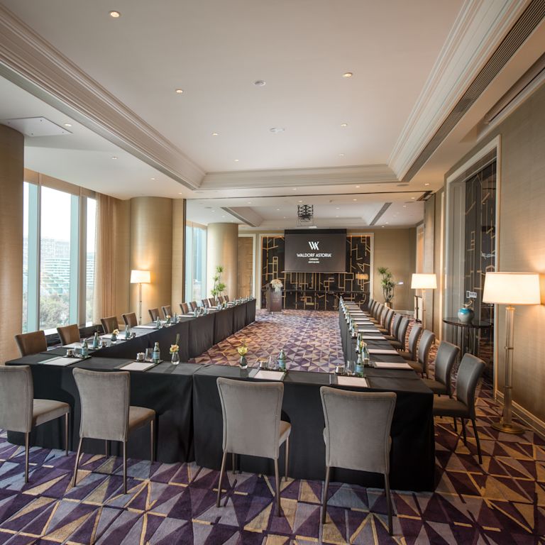 Meeting room with long U-shaped tables and chairs facing presentation screen, and floor-to-ceiling windows with outdoor view