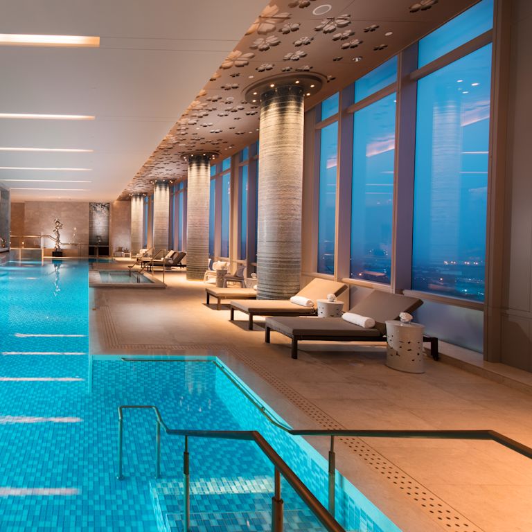 Lavish indoor pool with lounge chairs and floor-to-ceiling windows with outdoor view, lit up at night