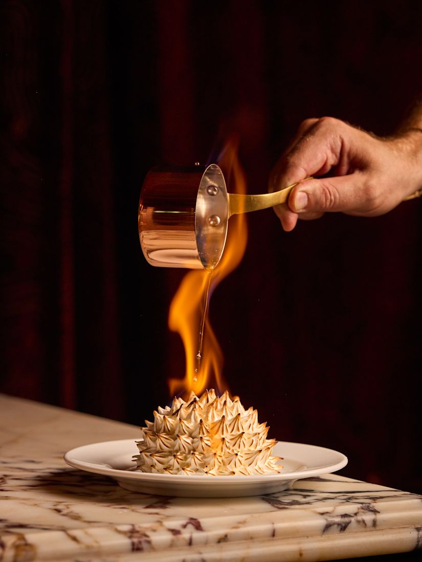 A hand pours flaming liquid from a copper ladle onto a baked Alaska dessert served on a plate over a marble table.