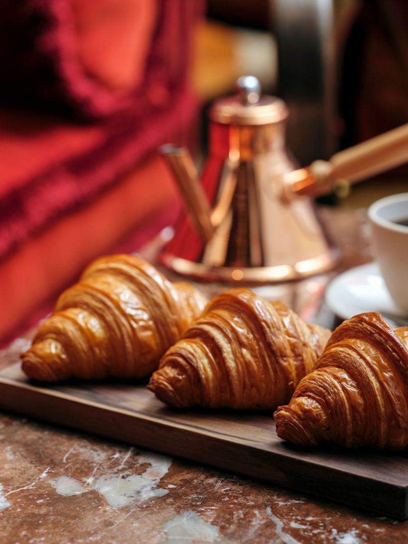 The Library breakfast with three croissants on a wooden board, a cup of coffee, and a copper kettle on a marble table.-previous-transition