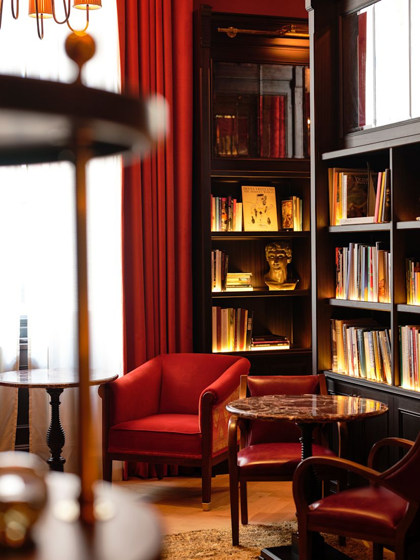 The Library with a cosy reading nook with red armchairs, a round table, dark wood bookshelves, and illuminated book spines.-next-transition