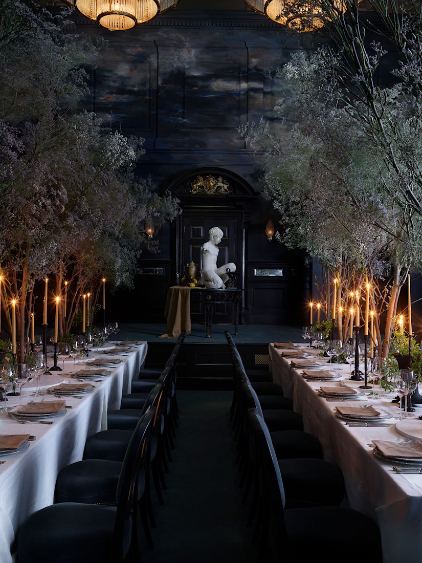 A grand dining room with two long tables, lit candles, tall branches, dark walls, and a stormy mural on the walls and ceiling.-next-transition