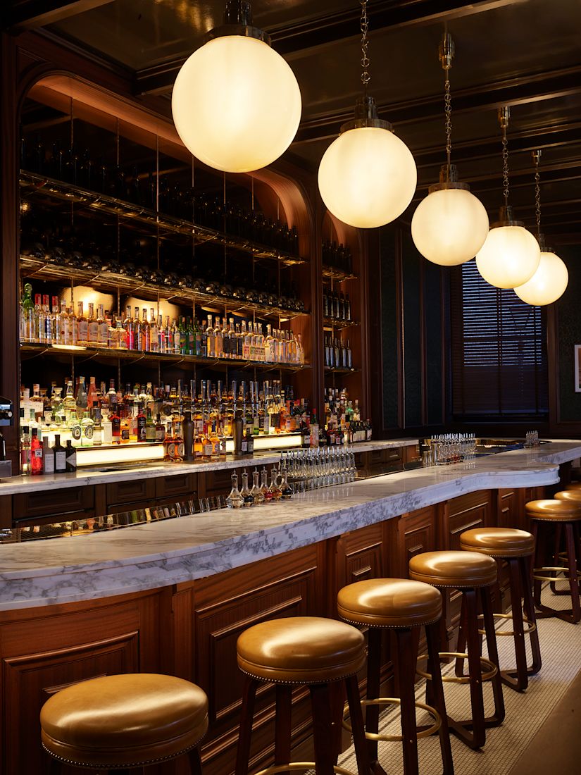 A bar with a marble countertop, wooden stools, shelves of liquor bottles, and hanging globe lights.-next-transition