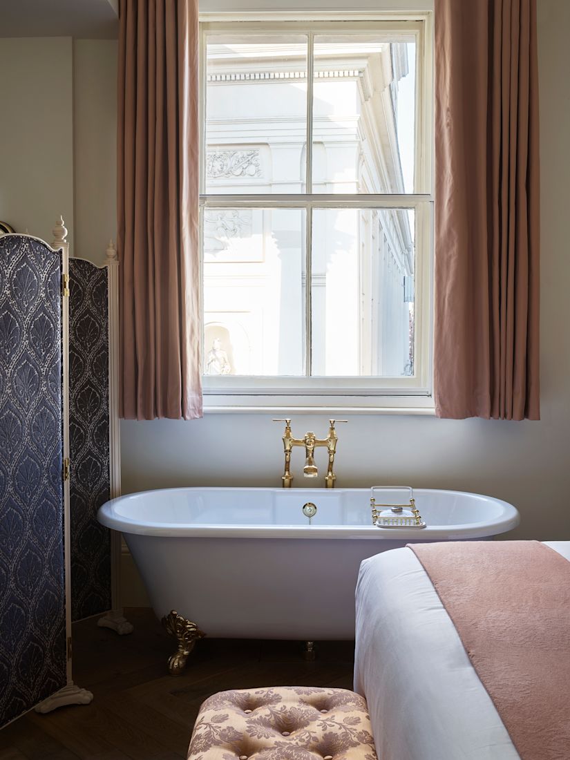 Bathtub with gold fixtures by a window with pink curtains and a black privacy screen.-previous-transition