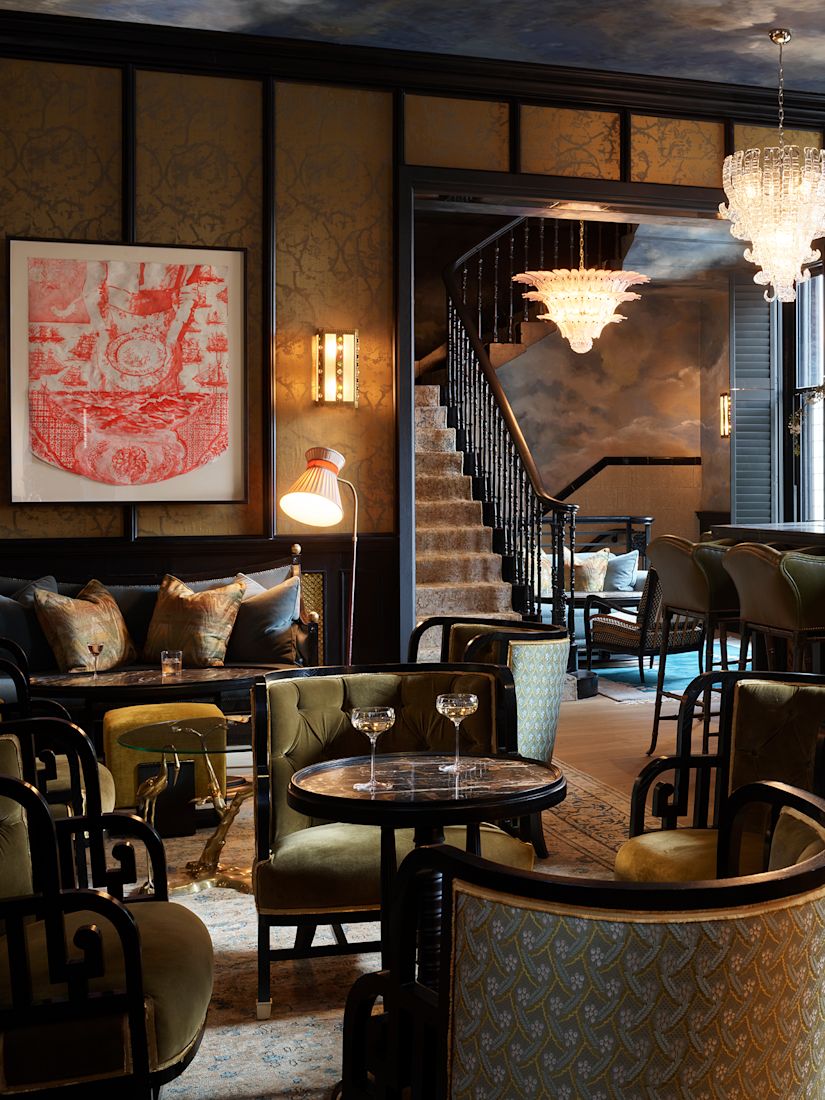 A vintage-style bar with green upholstered chairs, chandeliers, a staircase, and a art on the wall.-previous-transition