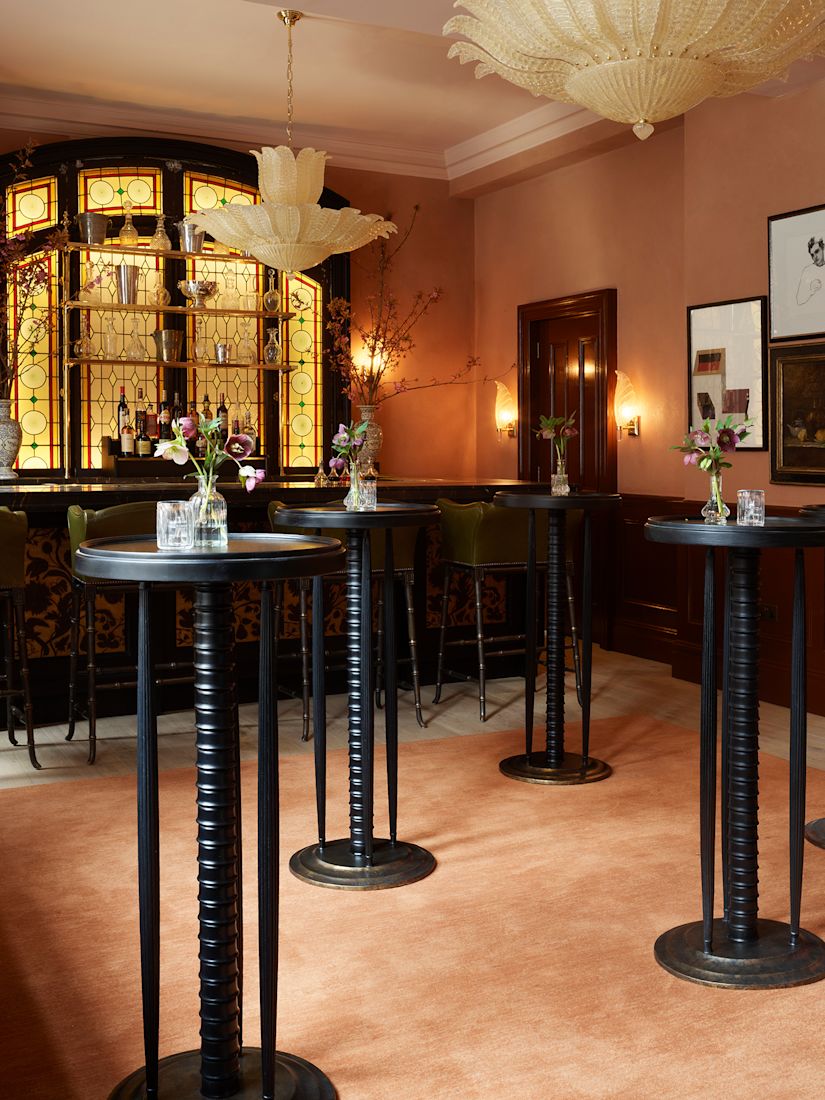 A bar area with tall black tables, a stained glass window, green bar stools, and floral arrangements on tables.-next-transition