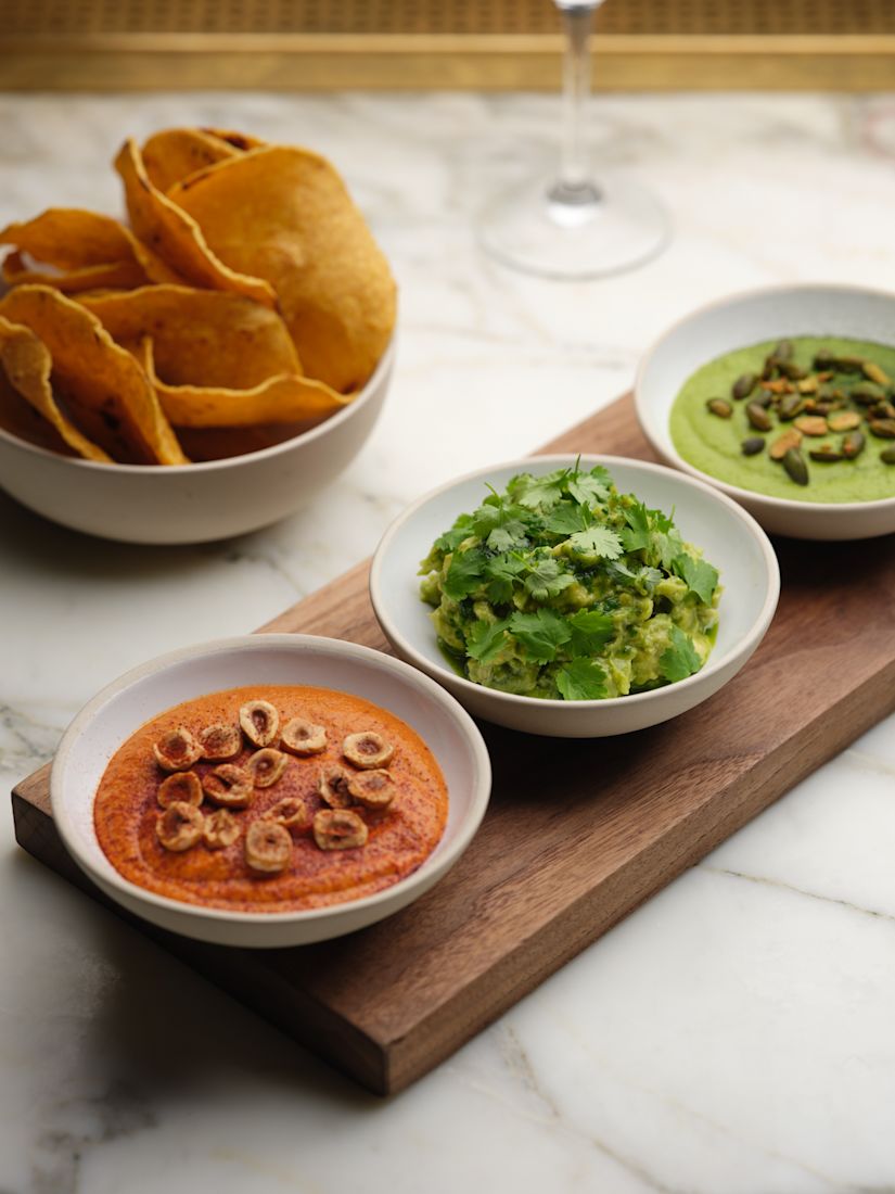 A wooden board with bowls of red dip topped with nuts, guacamole, and green salsa with seeds, next to a bowl of chips-next-transition