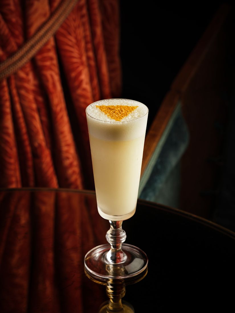 A cocktail tall glass of frothy yellow drink with a triangular powdered pattern on top.-next-transition