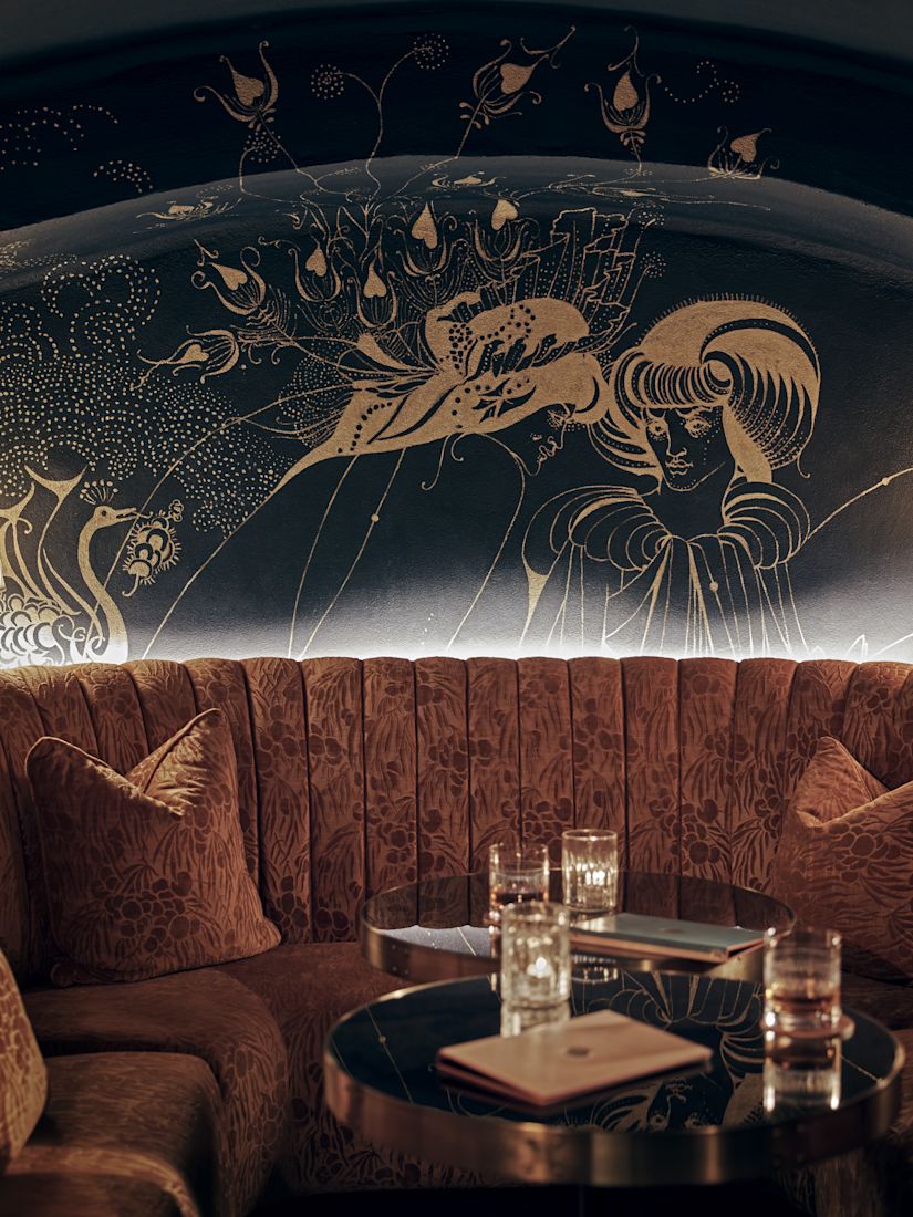 A plush curved sofa with patterned fabric, glass tables, and a wall mural of intricate gold designs.-previous-transition