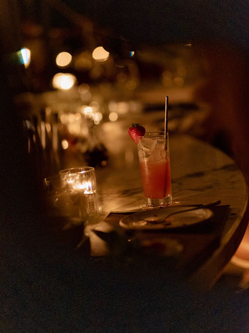 Cocktail in a tall glass with a strawberry garnish beside a candle on a dimly lit bar table-previous-transition