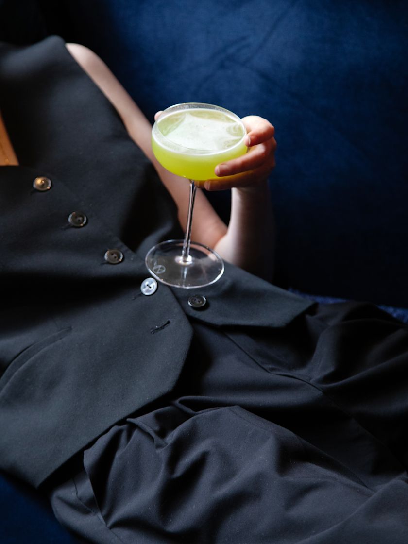 Person in dark clothing reclining and holding a bright green cocktail in a coupe glass.-next-transition