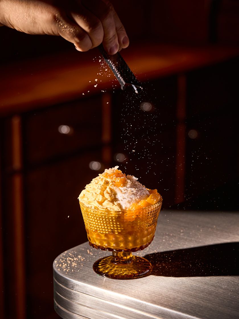 A hand grates garnish over a dessert served in an amber glass goblet on a metal-rimmed counter.-previous-transition