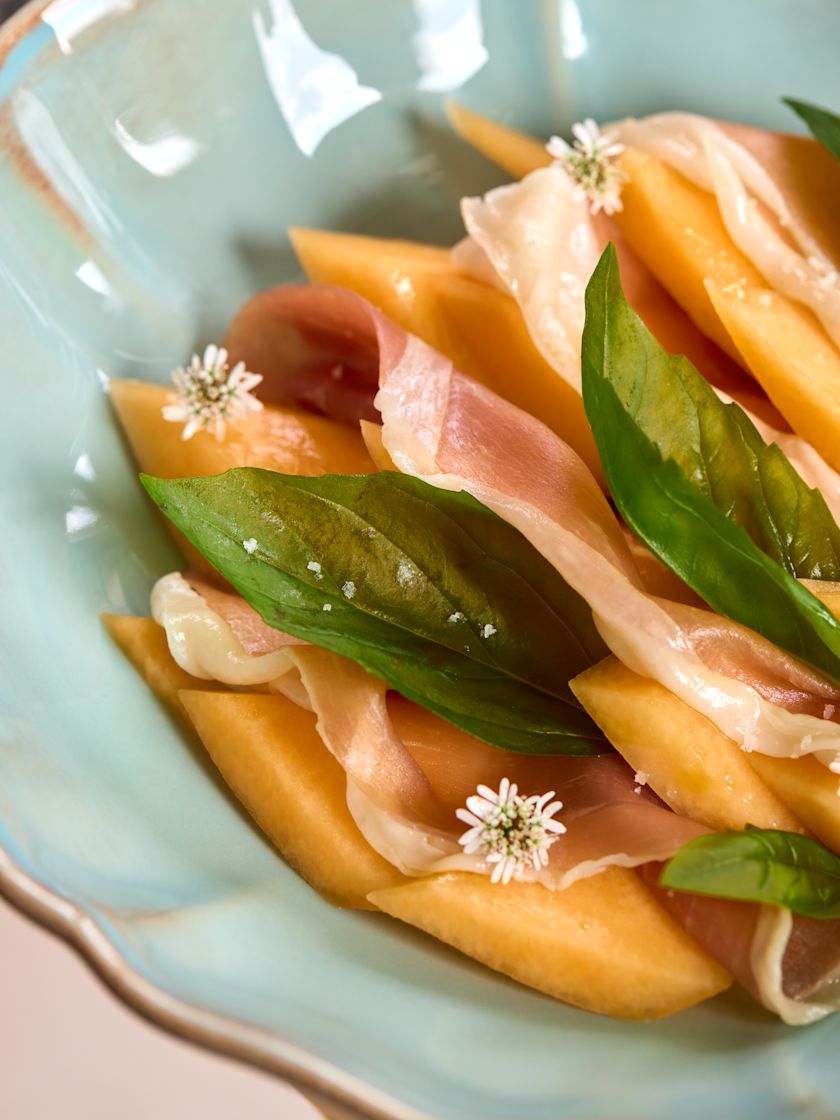 A light blue bowl filled with sliced melon, prosciutto, basil leaves, and small white edible flowers.-next-transition