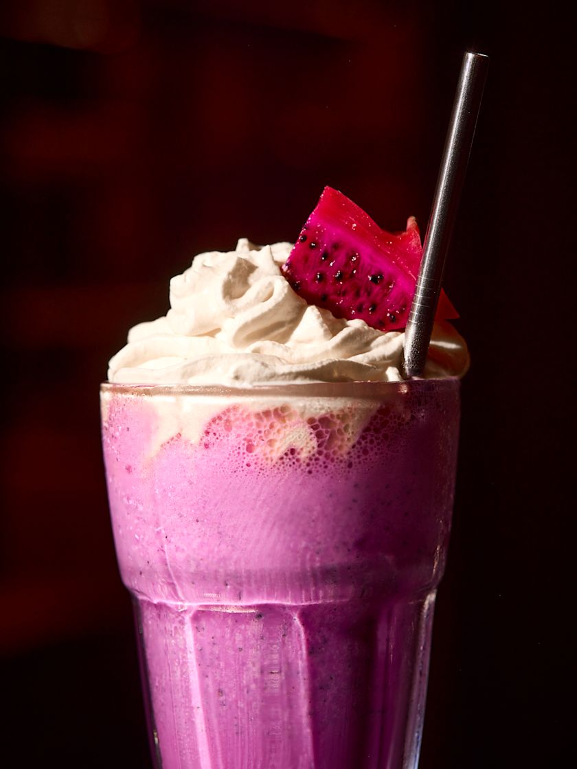 A tall glass of pink dragon fruit milkshake topped with whipped cream, a slice of dragon fruit, and a metal straw.-next-transition