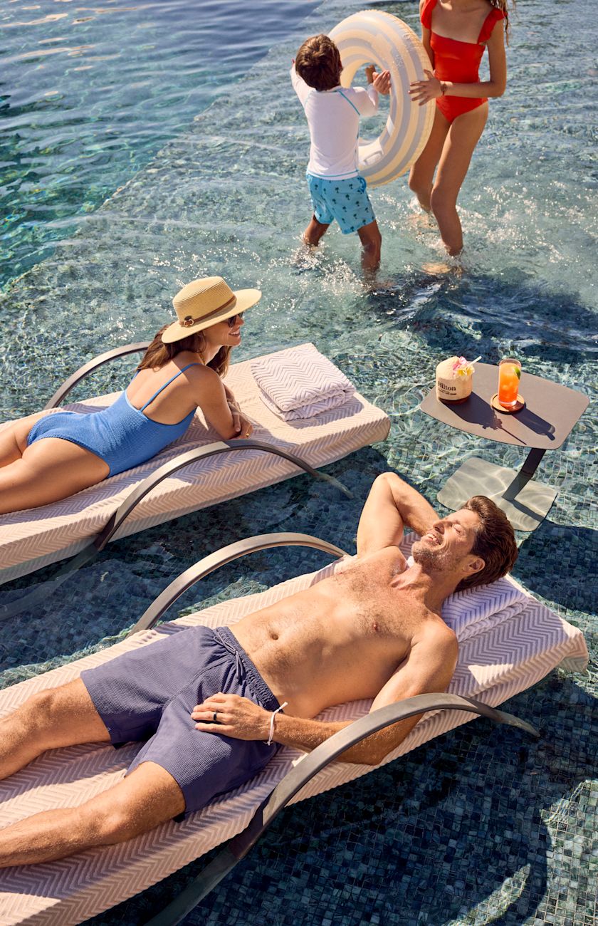 Ayah dan ibu bersantai di kursi santai di kolam renang sementara putra dan putri mereka terpercik di kolam renang