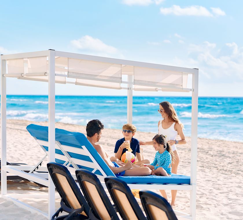 Family on the beach sitting on and standing around a large covered multi-person lounge seat.