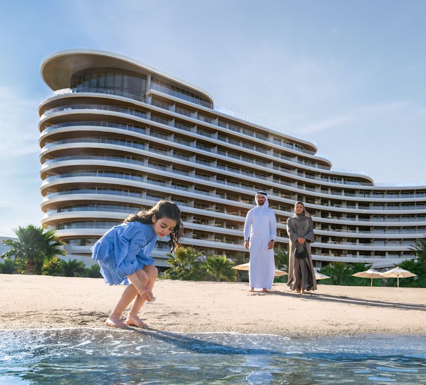 Man and woman watching young girl play on beach, with hotel exterior behind