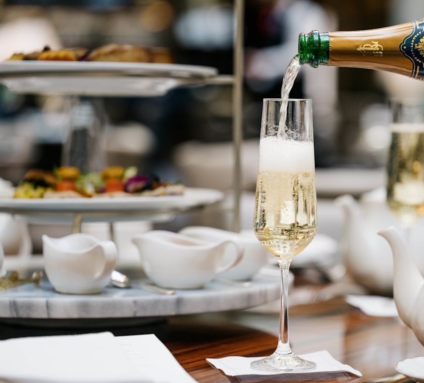 Șampanie turnată într-un pahar cu ceai de după-amiază în fundal