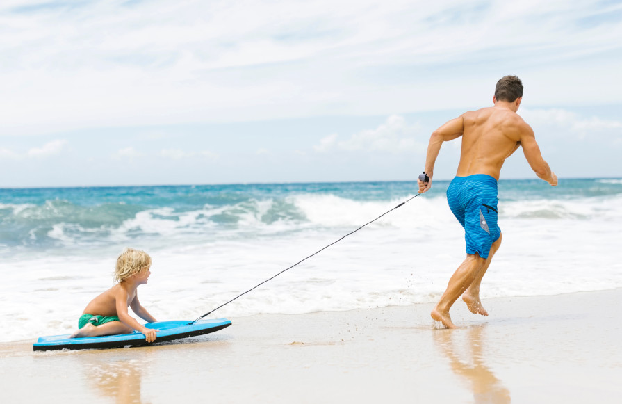 Man pulling child on surf board along beach