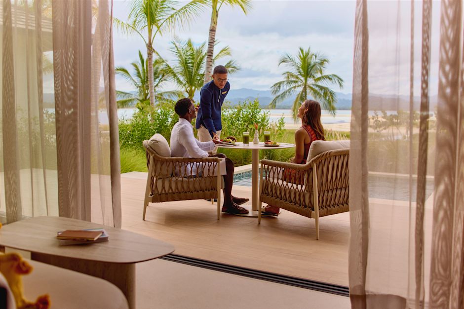 Two people dining on a shaded outdoor terrace while a server places food on the table, with views of palm trees, a pool, and distant water.