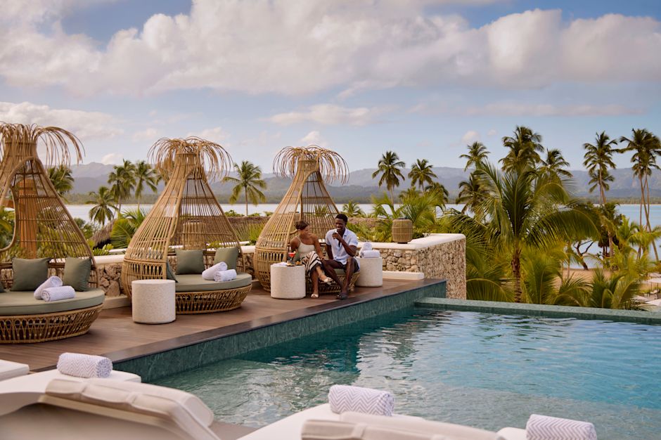 Two people relaxing beside an elevated pool on woven lounge pods, surrounded by palm trees and views of the ocean and distant hills under partly cloudy skies.