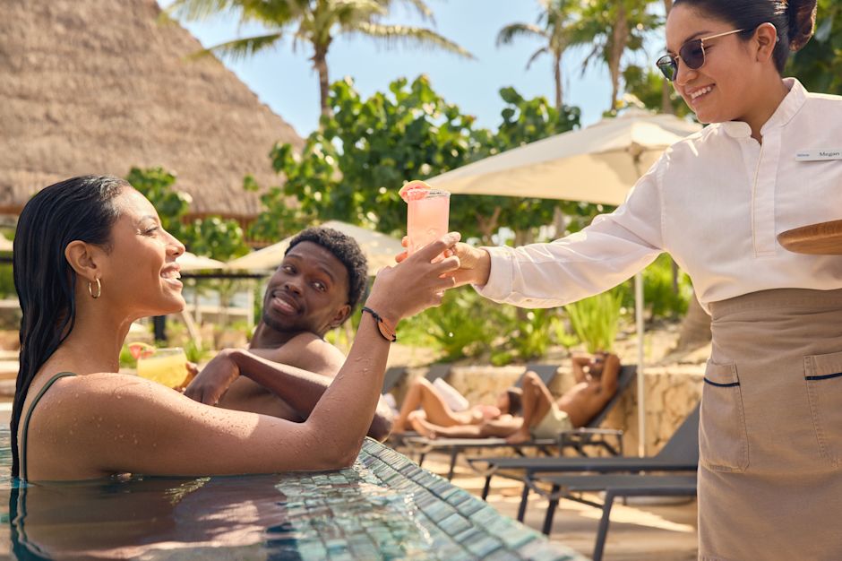 Vrouw en man loungen in het zwembad terwijl een teamlid de vrouw een verfrissend drankje serveert.