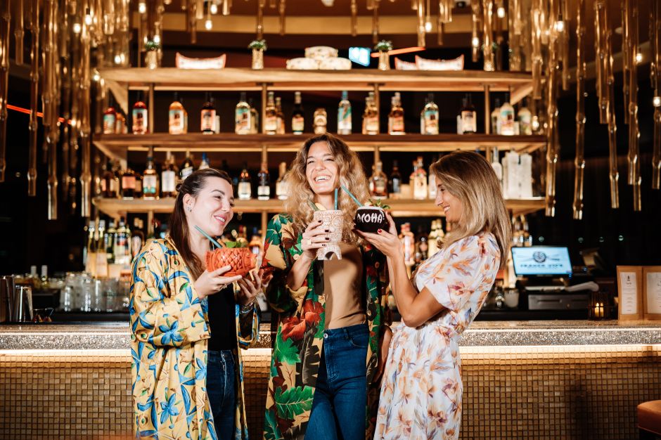 Three woman smiling and holding drinks in front of bar