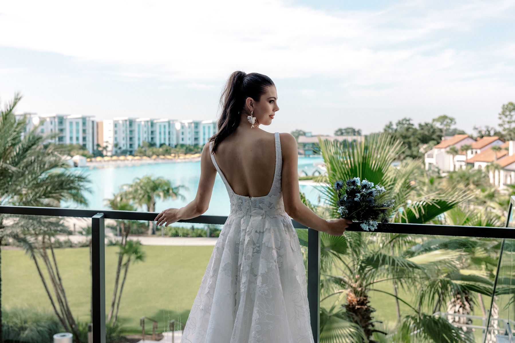 Mariage dans une robe fleurie tenant un bouquet sur un balcon surplombant l'eau, les palmiers et les bâtiments du complexe.