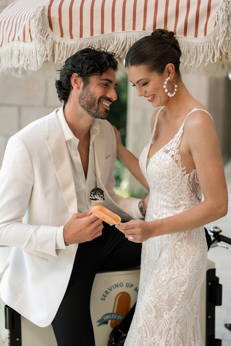 Couple in wedding attire smiling together outdoors near stone wall and greenery.