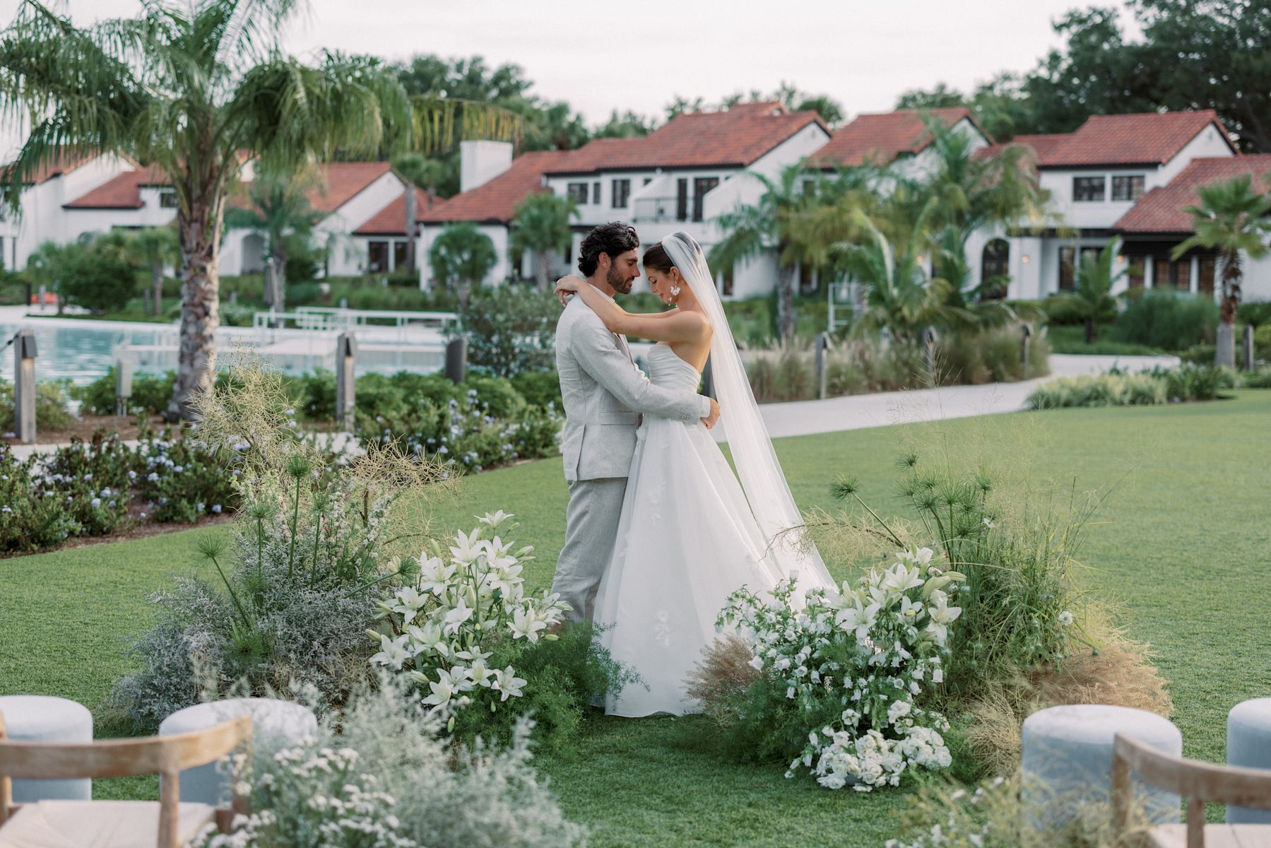Les mariés s'embrassent dans un jardin avec une voûte florale blanche et des palmiers en arrière-plan.