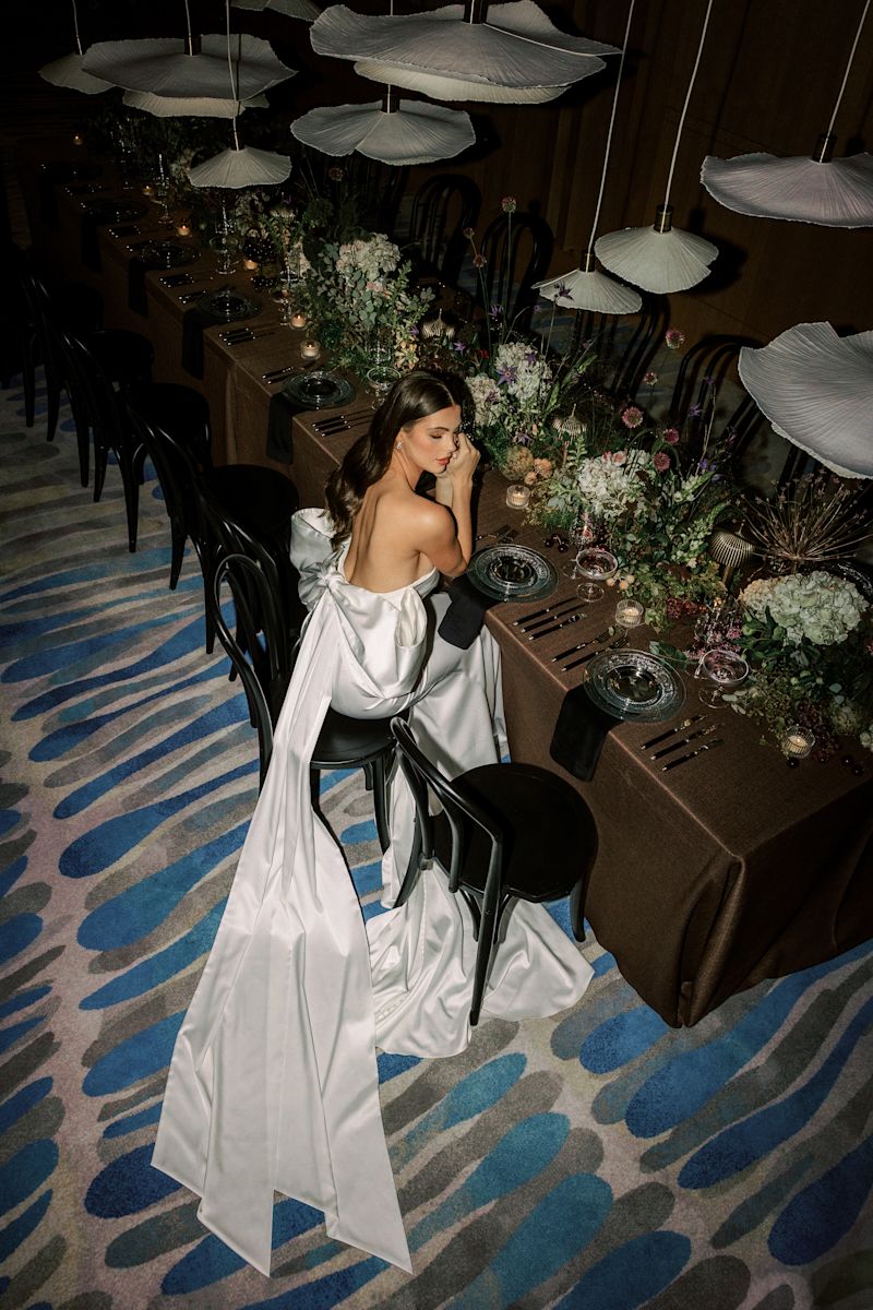 Mariée dans sa robe de mariée assise sur une chaise à une table formelle avec de la végétation.