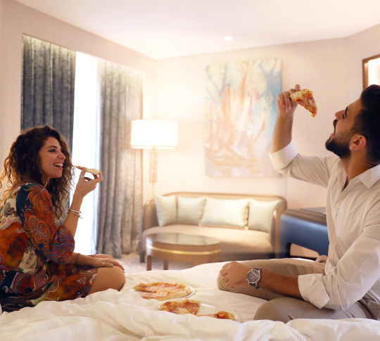 A couple eating pizza and laughing on the bed of the hotel room.