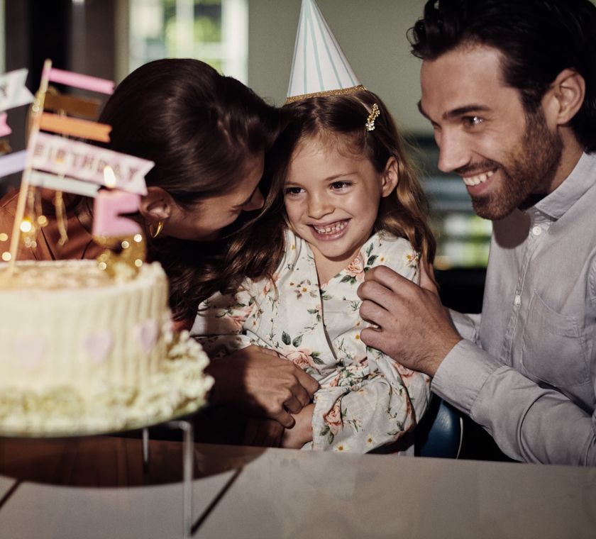 A mother and father at either side of their daughter who is smiling at a birthday cake.